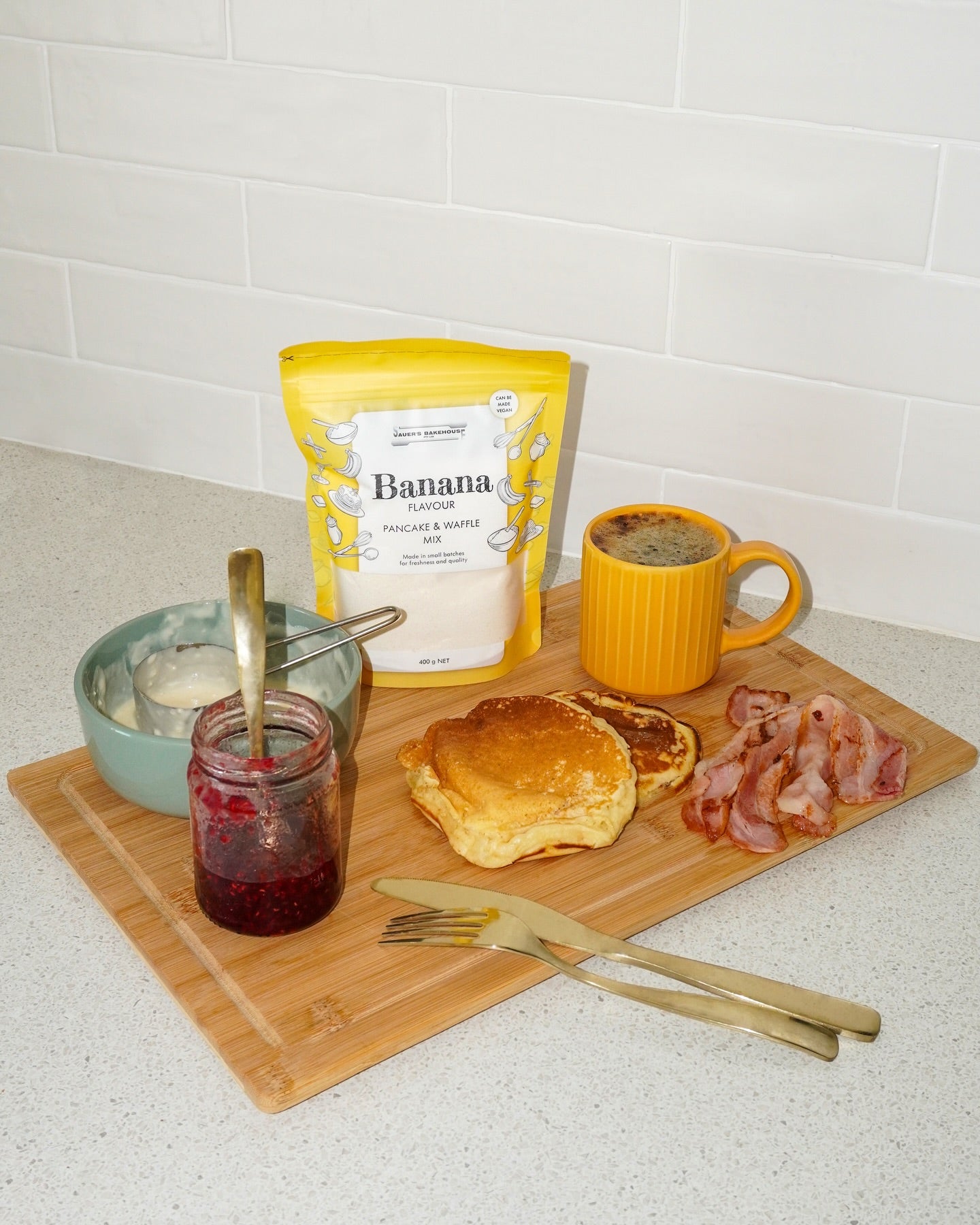 Breakfast setup with pancakes, bacon, jam, and a bag of banana flour on a wooden utting board.