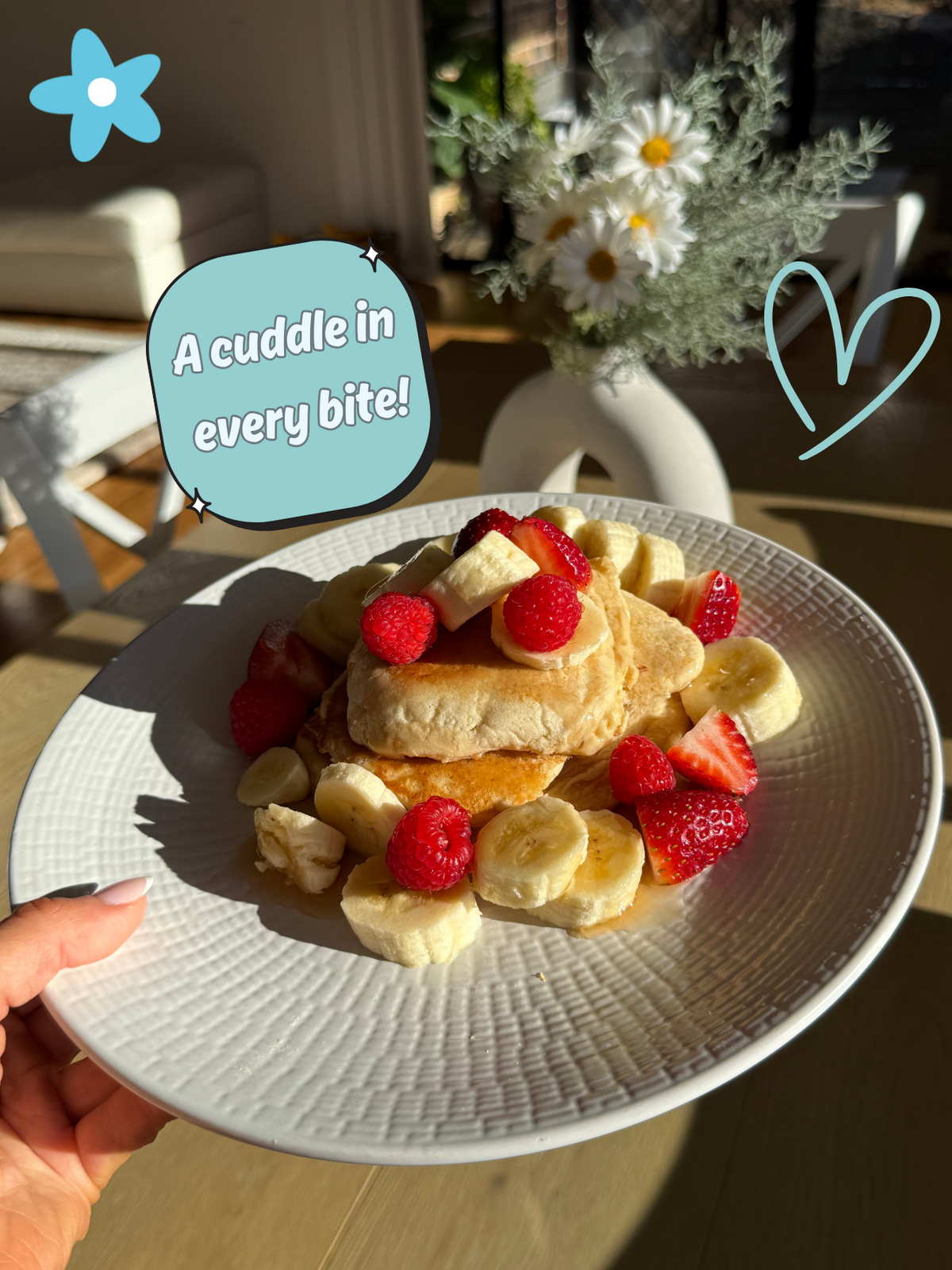 Plate of pancakes with fruit on a table, with text overlay 'A cuddle in every bite!' - Sauer's Bakehouse